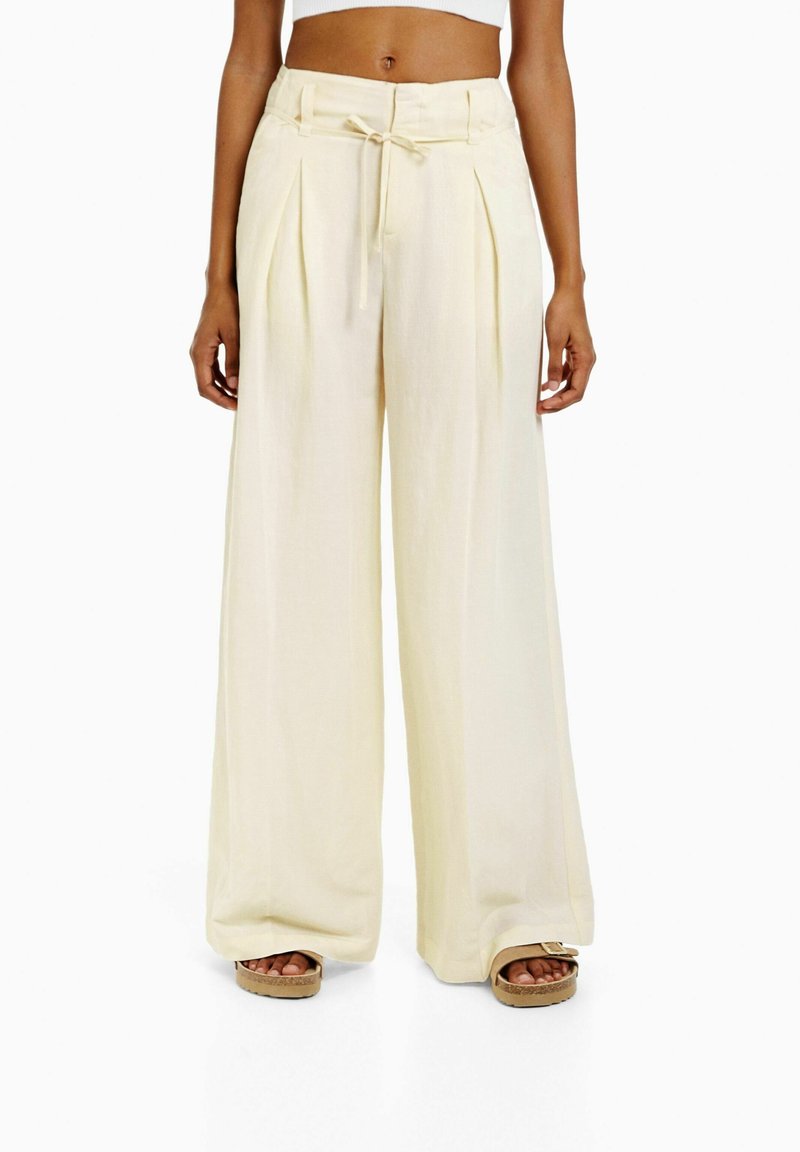Krämfärgade vida byxor med dragsko i midjan, plisserad design och mjuk textur, tillsammans med bruna plattformssandaler.