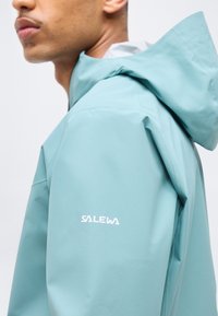 Κλείσιμο σε άτομο που φοράει γαλάζιο μπουφάν με κουκούλα, με το όνομα της μάρκας "Salewa" τυπωμένο στον πάνω βραχίονα.