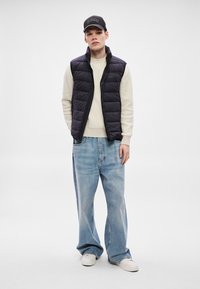 Melna pūkaina veste pār krēmkrāsas ribotu džemperi, kombinēta ar gaiši zilām platajam kāju džinsām un baltiem sporta apaviem. Modelis valkā melnu cepi.