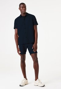 Camisa de manga corta, de color azul marino oscuro, con botones en la parte delantera y cuello, combinada con pantalones cortos a juego. Usada con zapatillas deportivas de color claro.