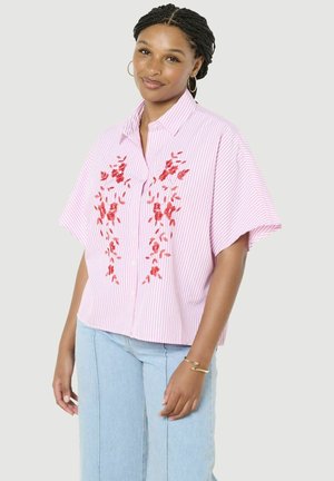 Kobieta w różowo-białej koszulce w paski z czerwonym kwiatowym haftem i jasnoniebieskich dżinsach, stojąca na jasnym tle.