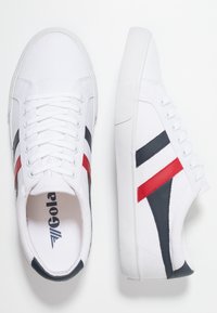 Sneakers bianchi in tela con accenti di design neri e rossi, lacci piatti, suola in gomma texturizzata e logo sulla soletta.