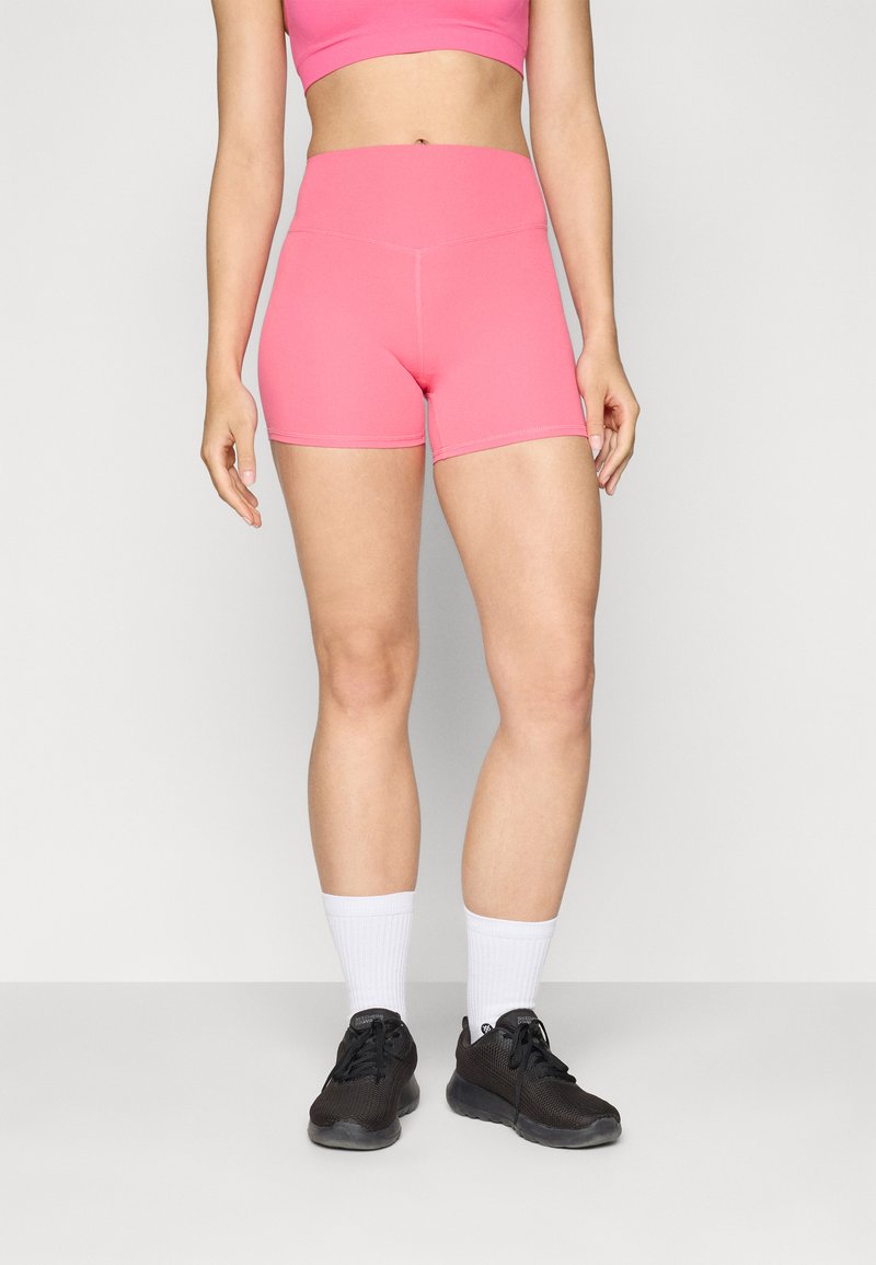 Rosa treningsshorts med høy midje, tettsittende design og kontrasterende sømmer. Brukt med svarte joggesko og hvite ankelsokker.