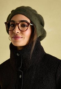 Czarna, teksturowana kurtka sherpa z wysokim kołnierzem i zapięciem na guziki; zielona rypsowa czapka; okrągłe okulary w kolorze tortoishell; neutralne tło.