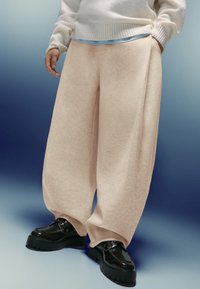 Osoba oblečená v béžových širokých nohaviciach, čiernych robustných šnurovacích topánkach, krémovom svetri a svetlomodrej košeli pod ním.