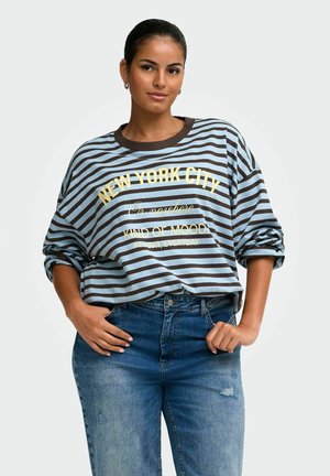 Femme aux cheveux foncés portant un pull rayé bleu et marron avec un texte, légèrement penchée, portant un jean bleu avec les mains près des poches sur fond blanc.