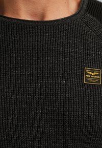 Close-up van een zwart geribd truien met een gele en zwarte PME Legend patch op de borst met de tekst "American Classic Tested at High Altitude."