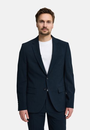 Mężczyzna o jasnobrązowych włosach i brodzie, ubrany w ciemnogranatową marynarkę na białej koszulce z okrągłym dekoltem, na jednolitym białym tle.