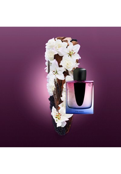 Perfume de Shiseido en un frasco de vidrio degradado, con una tapa negra. Rodeado de flores blancas y detalles en madera oscura sobre un fondo púrpura.