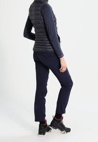 Maritime Outdoor-Hose, blaues Langarmoberteil, gepolsterte schwarze Weste und schwarze Wanderschuhe mit roten Akzenten, mit einem Blumenlogo auf der Hose.