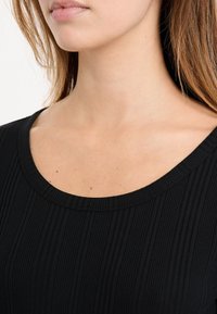 Zbliżenie na szyję i obojczyk kobiety noszącej czarny, prążkowany top z dekoltem, z prostymi brązowymi włosami.