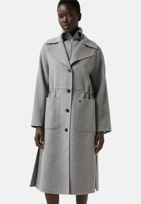 Kvinde iført en lang grå uldfrakke med brede revers, fire frontknapper, store påsatte lommer og sideslidsser, over en lynlås-sweater.