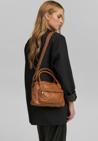 Borsa a mano in pelle color cuoio con manici superiori doppi e una tracolla removibile. Presenta una tasca con zip sul davanti e una texture liscia.