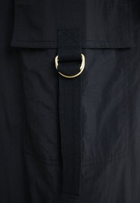 Tissu noir avec une finition mate, présentant un rabat plié sécurisé par un anneau en métal doré et une sangle noire texturée en dessous.