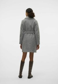 Manteau gris texturé avec une taille ceinturée, un col surdimensionné et des poches latérales, porté avec de grandes bottes en cuir marron.