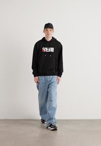 Svart hettegenser med hvit og rød logo, lyseblå vide jeans og svarte sneakers, båret med en svart caps.
