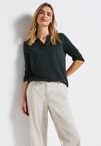 Tummanvihreä, kolmannelta neljännekseltä hihoilla varustettu paita, jossa on V-pääntie ja röyhelöhihojen päät, yhdistettynä vaaleanbeigeihin, löysiin housuihin. Sujuva kangas.