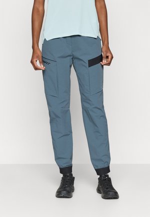 Grå træningsbukser med tætsiddende ben, elastiske manchetter og flere lommer. Har sorte accenter og et logo på venstre lår.