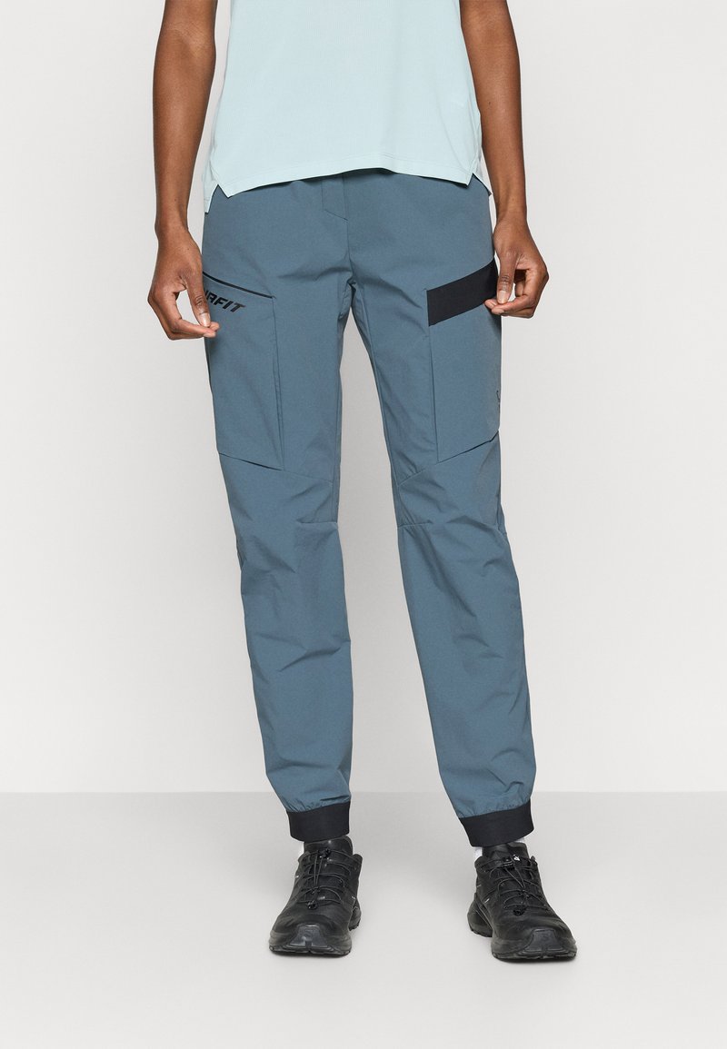 Γκρι αθλητικά παντελόνια με σφηνοειδή ποδιά, ελαστικές μανσέτες και πολλαπλές τσέπες. Διαθέτουν μαύρες λεπτομέρειες και λογότυπο στον αριστερό μηρό.