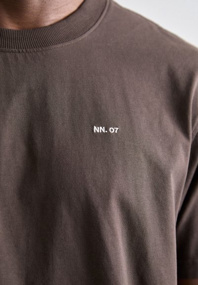 T-shirt en coton marron avec un col rond côtelé et un petit logo "NN. 07" brodé sur la poitrine. Tissu doux et texturé.
