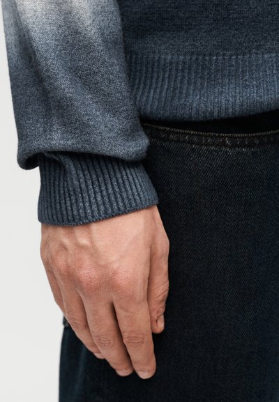 Γκρι πλεκτό πουλόβερ με σχεδιασμό διαβάθμισης, μανσέτες με ριμπ ύφανση και λεία υφή, σε συνδυασμό με σκούρο τζην τζιν, που αναδεικνύει περίτεχνη ραφή.