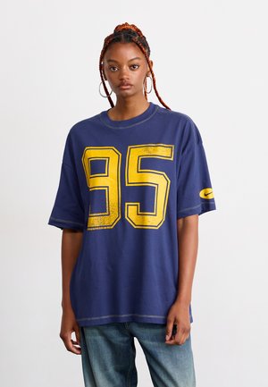 Young person with braided hair wearing large hoop earrings, a loose navy shirt with yellow number 95, and blue jeans, standing against white background.
