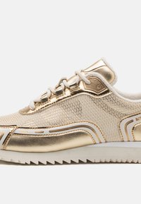 Scarpe da ginnastica dorate con tomaia in rete, dettagli metallici e lacci bianchi. Presentano un design testurizzato e una suola in gomma seghettata.