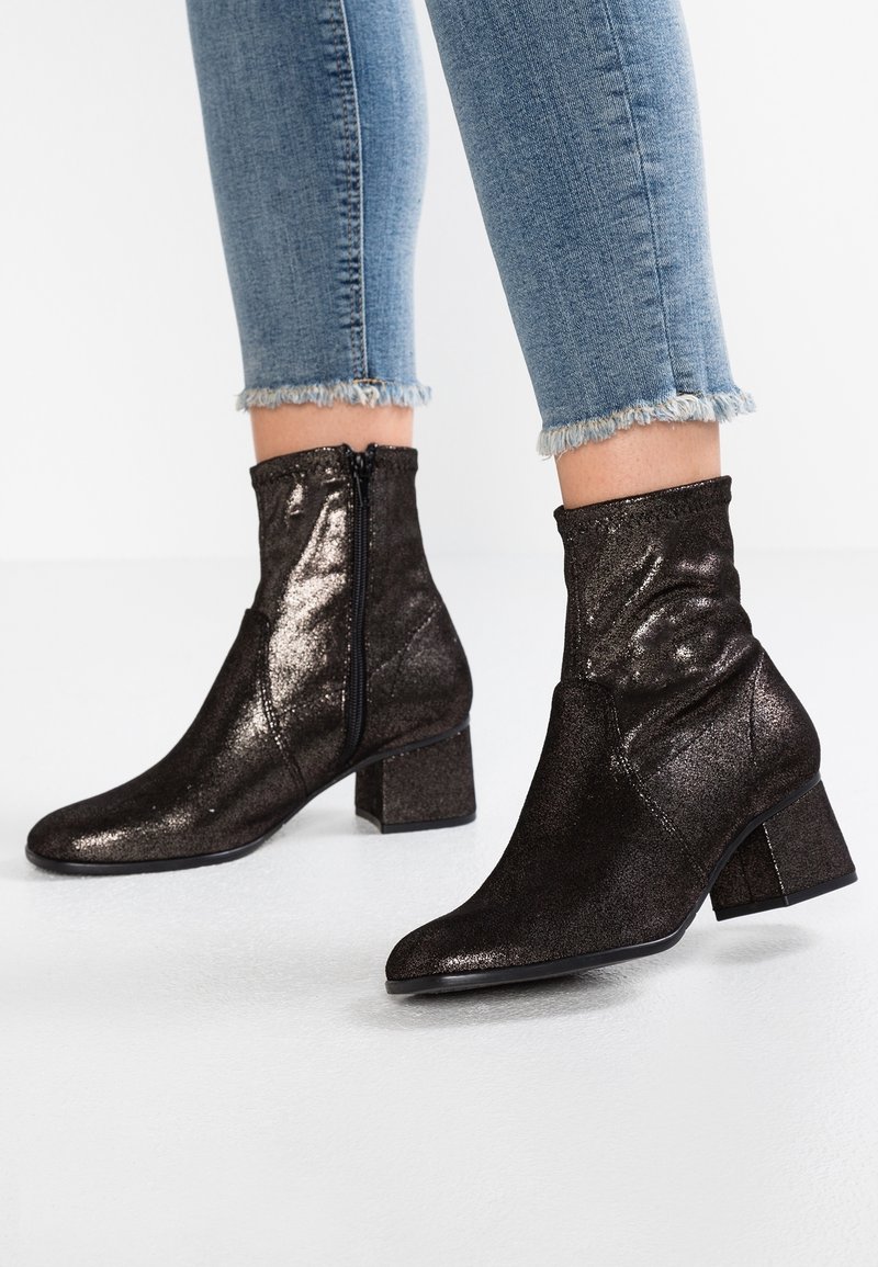 Personne portant des bottines noires étincelantes à talons blocs et fermetures éclair latérales, associées à un jean skinny bleu clair à ourlet effiloché.