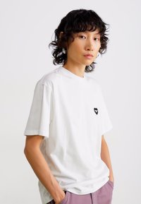 Jeune personne avec des cheveux foncés et bouclés, portant un t-shirt blanc avec un petit emblème en forme de cœur noir et un pantalon lilas clair, se tenant les mains dans les poches.