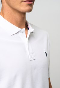 Polo blanc en tissu texturé, avec un col boutonné et un petit logo noir sur la poitrine gauche. Détails de contraste subtils à l'intérieur du col.