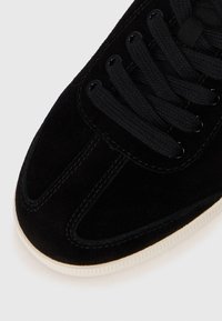Baskets en suède noir avec une tige texturée, dotées de lacets plats noirs et d'œillets ronds noirs. Semelle en caoutchouc blanc avec texture striée.