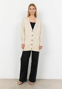 Cardigan beige con scollo a V, bottoni davanti e due tasche, abbinato a pantaloni neri a gamba larga e sandali con motivo.