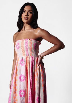 Vrouw draagt een strapless, vloeiende jurk met roze en oranje geometrische patronen, poseert met één hand op haar heup tegen een effen achtergrond.
