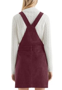 Burgunderfarbenes Cordkleid mit verstellbaren Kreuzträgern, tailliertem Schnitt und strukturiertem Oberflächenmaterial. Kombiniert mit einem cremefarbenen, kabelgestrickten Pullover.