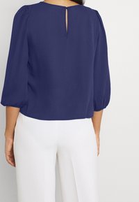 Blouse bleu marine avec des manches bouffantes trois-quarts. Dotée d'un détail en forme de trou de serrure à l'arrière et fabriquée dans un tissu lisse et léger. Associée à un pantalon blanc.