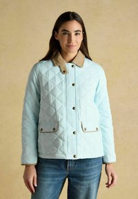 Mujer con cabello largo y oscuro, vistiendo una chaqueta acolchada azul claro con cuello y bolsillos abotonados en tono marrón claro, de pie frente a un fondo beige.