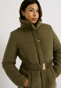 Abrigo acolchado color verde oliva con cuello alto, cintura con cinturón y botones de presión. Presenta secciones acolchadas y bolsillos laterales. Textura suave.