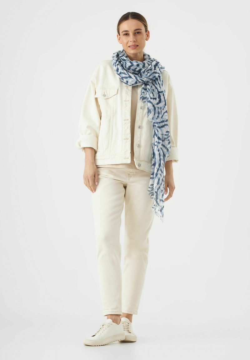 Écharpe légère bleu et blanc à motifs drapée autour d'un modèle, associée à une veste en denim crème, un haut beige et un jean clair.