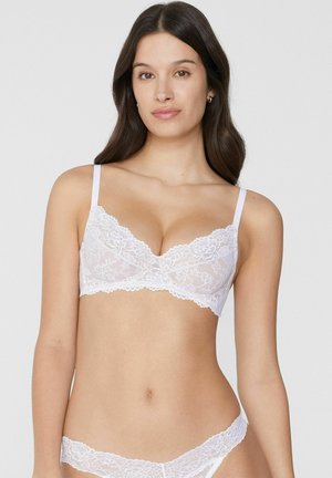 Frau trägt weißen Spitzen-BH und passende Spitzen-Unterhose, steht vor einem schlichten hellgrauen Hintergrund.