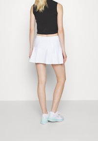 Falda blanca plisada con una cinturilla ancha, combinada con un top negro sin mangas y zapatillas multicolores, vista desde atrás.