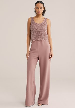 Jeune femme portant un haut sans manches mauve avec des détails en perles et un pantalon large assorti, posant devant un fond beige uni.