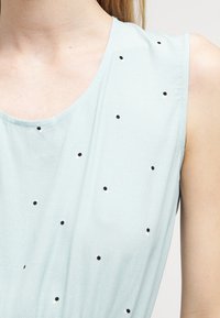 Vaaleansininen hihaton paita, joka on valmistettu pehmeästä kankaasta ja siinä on musta-valkoinen pilkku-kuviointi sekä pyöreä pääntie.