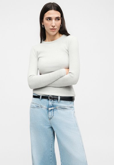 Γυναίκα με μακριά σκουρόχρωμα μαλλιά, φοράει ασημένια κρίκους στα αυτιά, ανοιχτό γκρι ριγωτό μακρυμάνικο τοπ, ανοιχτό μπλε τζιν και μαύρη ζώνη, με σταυρωμένα τα χέρια.