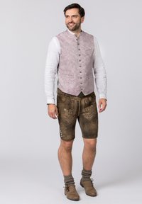 Traditionelles bayerisches Outfit mit einer gemusterten pinken Weste, braunen Lederhosen mit aufwendiger Stickerei und braunen Schuhen mit knielangen Socken.
