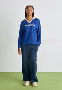 Ilgi piedurkņu zils krekls ar krēmkrāsas apkakli un "new balance" tekstu, apvienots ar tumši zilām biksēm un gaišām kedām. Strukturēta auduma.