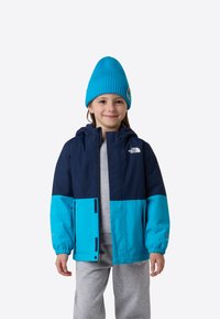 Jachetă cu blocuri de culoare albastru și bleumarin, cu glugă, realizată din material sintetic. Asortată cu o cagulă tricotată albastră și pantaloni casual gri.