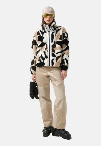 Imitatiebontjas met zwart-witte en beige abstracte patronen, gecombineerd met beige waterdichte broek en zwarte sneeuwlaarzen, handschoenen vasthoudend.