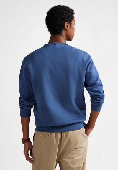 Zils kokvilnas džemperis ar ribveida aprocēm un apakšmalu, apaļa kakla izgriezumu un brīvu piegriezumu. Viegls tekstūra, nav rakstu vai rotājumu.