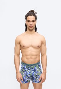 Homme athlétique avec dreadlocks portant un boxer vert à grand motif floral bleu, debout devant un fond clair uni.