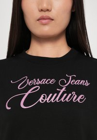 Juodas trumpomis rankovėmis marškinėliai, kurių priekyje yra didelis rožinis blizgus užrašas "Versace Jeans Couture". Klasikinio apvalaus kaklo dizainas.